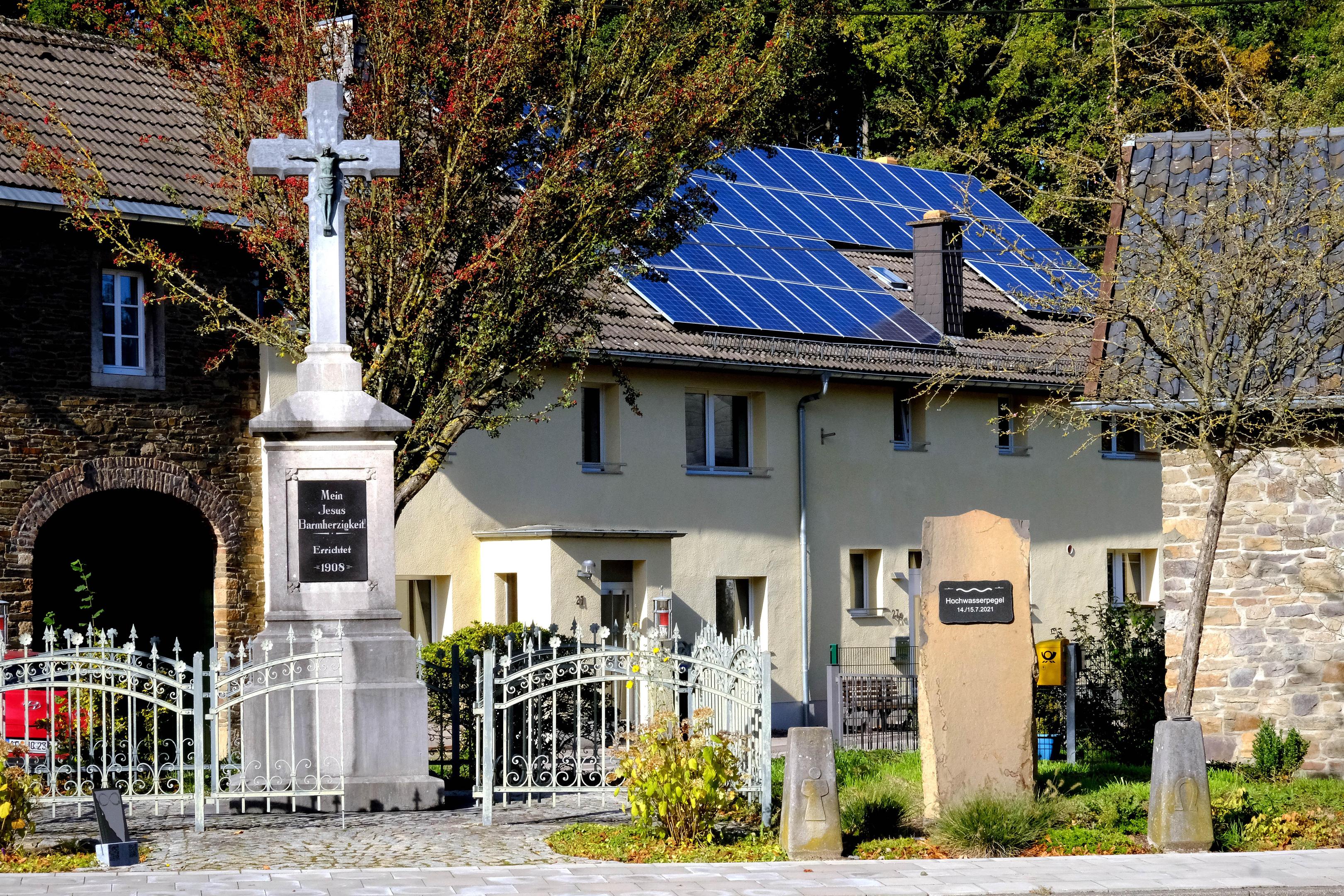 Das Missionskreuz ist der zentrale Blickfang auf dem Dorfplatz in Mulartshütte. Ein im Jahr 2023 errichteter Gedenkstein (re.) in direkter Nähe markiert den Pegel des verheerenden Hochwassers vom 14./15. Juli 2021. 